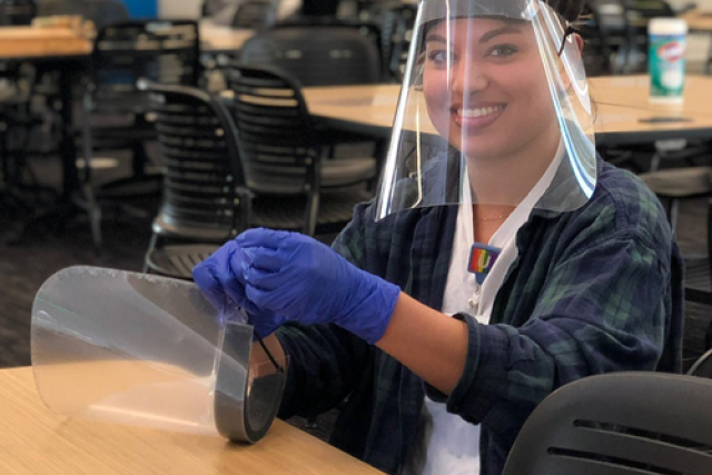 Smiling woman wearing gloves and a face shield assembles protective gear at a table in a socially distanced room.