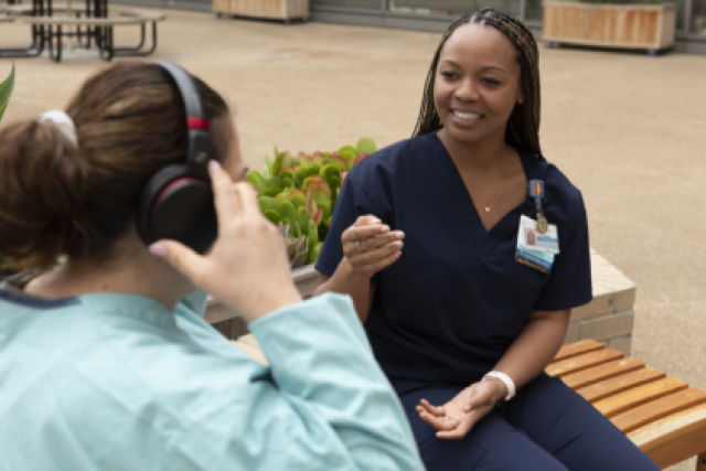  Shoni Taylor, wearing dark blue scrubs, smiles while talking to another woman with headphones on a bench outdoors.