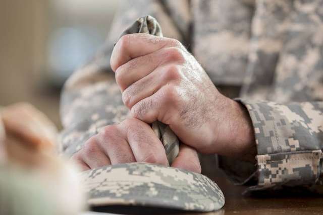 A close-up of a person in a military camouflage uniform whose hands are tightly clenched around fabric, resting on a table next to a camouflage cap.