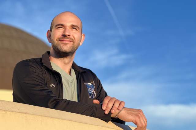 A doctor leans on a wall looking out into the distance, with the blue sky above.