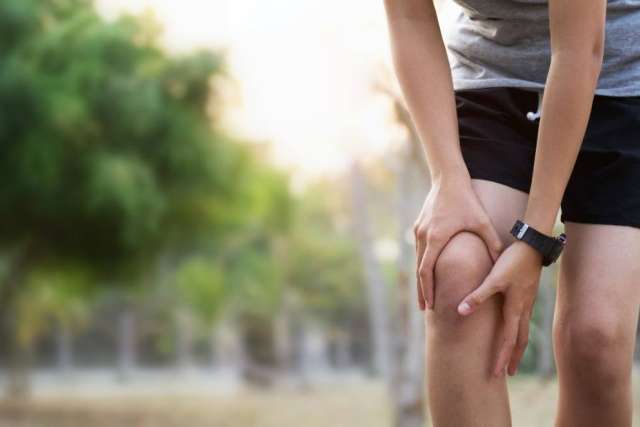 A person in shorts rubs their knee, indicating discomfort in a park setting.