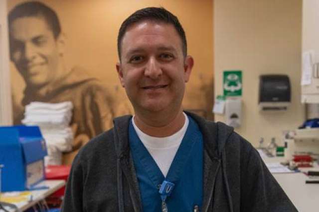 Person wearing blue scrubs and a dark jacket standing in a medical setting with supplies and equipment in the background.