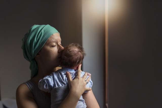 Woman in headscarf, fighting breast cancer while holding her newborn baby relaxing in cancer treatment hospital, patient standing next to hospital window. 