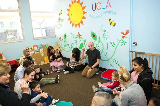A group of children and adults sit on the floor in a brightly colored room with a mural of the sun, plants, and bees on the wall.