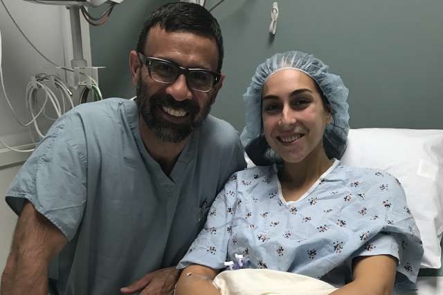 A patient in hospital attire sitting on a bed with a support person beside them.