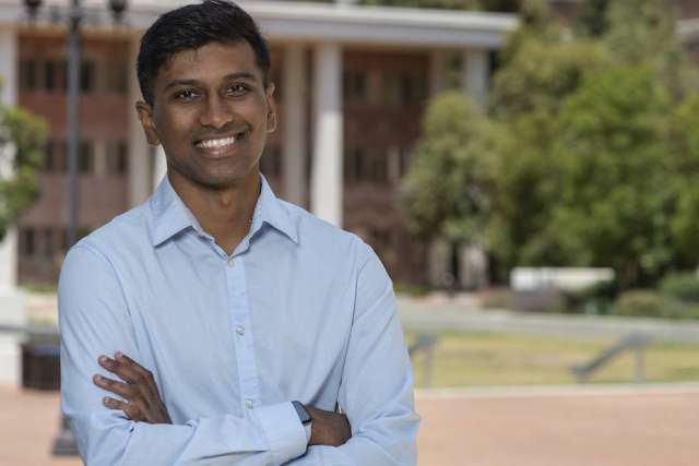 Vikram Krishna, a man with dark skin, short dark hair, and glasses, smiles confidently with his arms crossed outdoors.