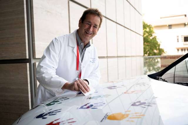 A smiling man in a white lab coat leans over to add a colorful handprint to the white hood of a car.