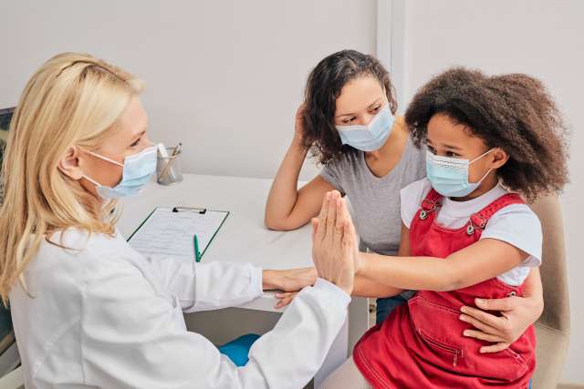 Masked female doctor high-fiving a masked child sitting on parent's lap in clinic.