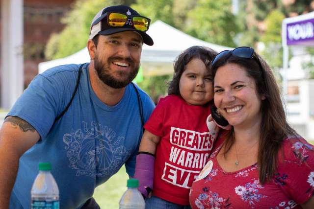 A smiling family outdoors: a bearded man in a blue shirt and baseball cap, a young child in a red "GREATEST WARRIOR EVER" shirt and cast, and a woman in a floral top.