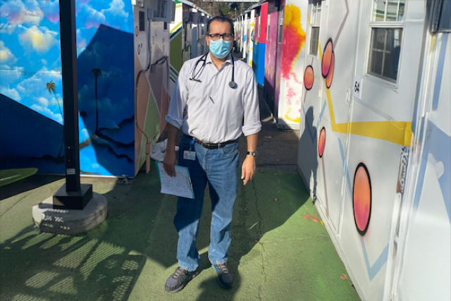 A healthcare worker wearing a mask and stethoscope stands in a colorful outdoor setting.