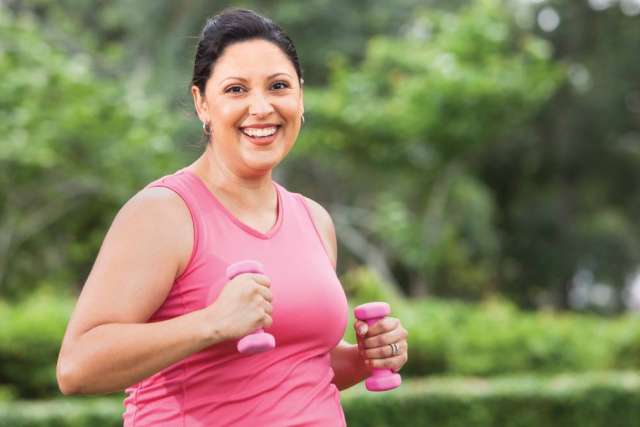  A smiling woman with dark hair pulled back wears a pink tank top while exercising outdoors, holding a small pink dumbbell in each hand.