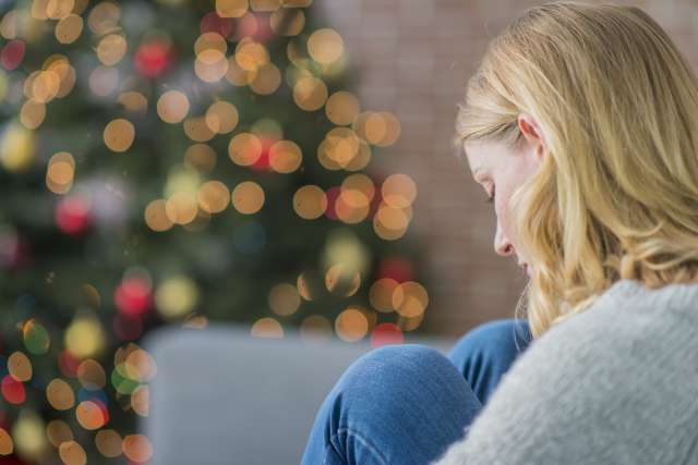A woman, sitting alone, looks said, with a Christmas tree in the background.