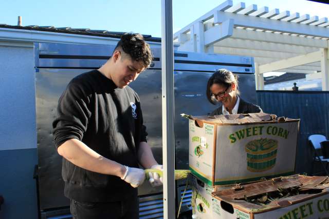 Two people preparing food with boxes of sweet corn outdoors.