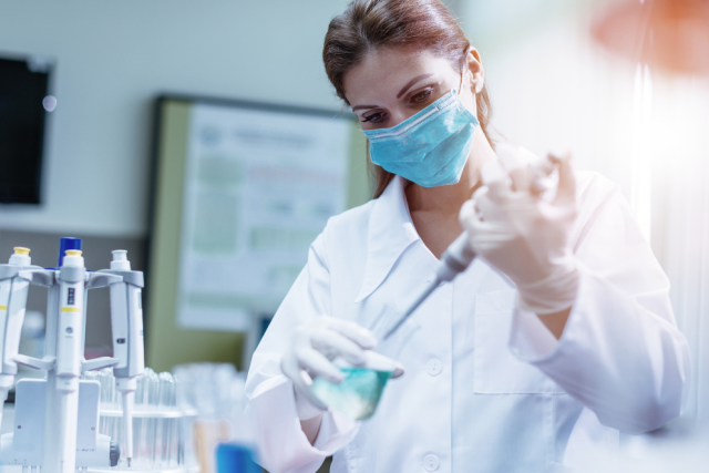  A masked and gloved scientist in a lab coat uses a pipette to transfer liquid in a brightly lit laboratory setting.