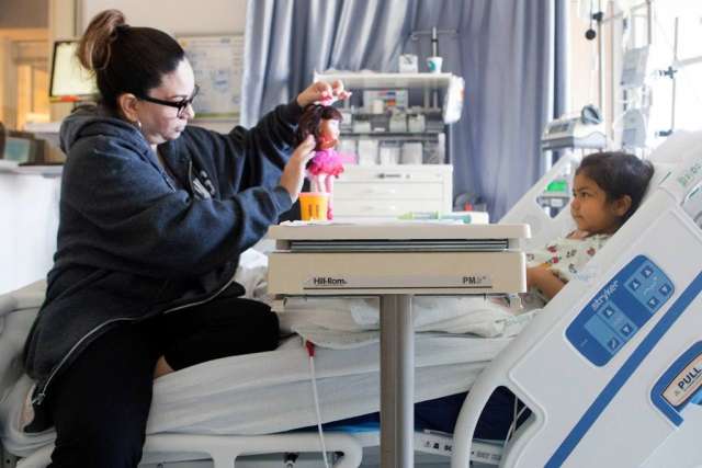A young girl, Giuliana, in a hospital bed with a woman sitting at her bedside, playing with a toy.