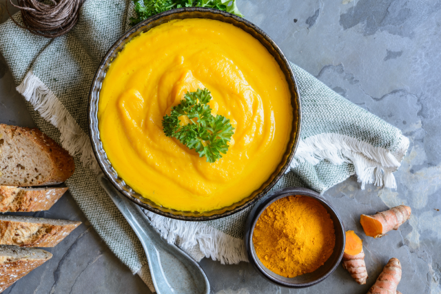 Creamy orange soup in bowl garnished with parsley; turmeric powder and roots nearby.