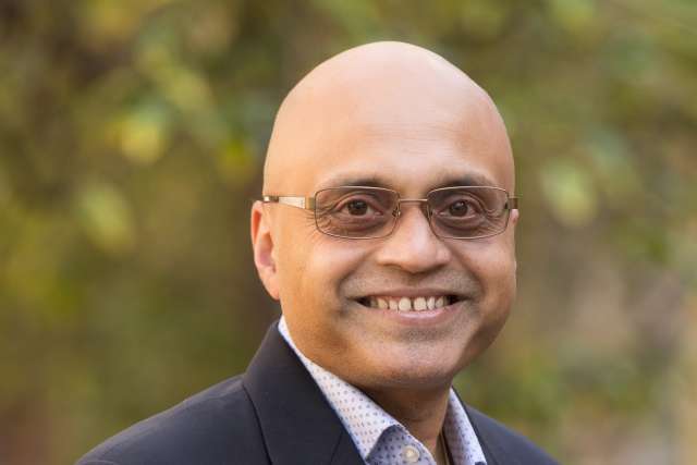 Headshot of person in dark suit and patterned shirt, outdoor background