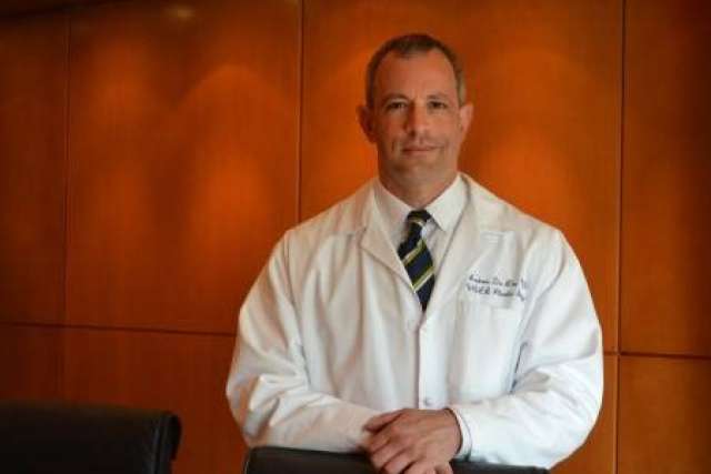 Dr. Andrew Da Lio with short hair and glasses wears a tie and a white lab coat, standing with his hands clasped on a chair.