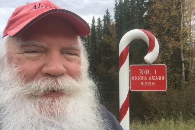 Man with white beard at No. 1 Santa Claus Lane sign shaped like a candy cane, standing in front of trees outdoors.