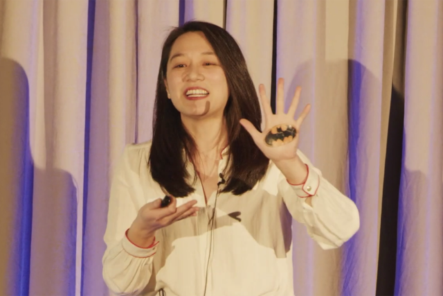 An-Chieh Feng smiles while giving a presentation. She holds up her left hand to reveal a small batman symbol painted on her palm.
