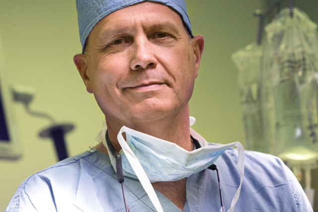 Dr. Glen Van Arsdell in scrubs and surgical cap stands confidently in an operating room, with IV bags and medical monitors behind him.