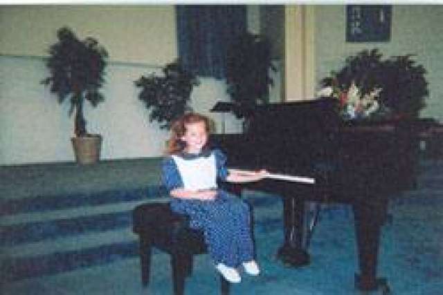 Child seated at a piano in a large room with blue carpeting and potted plants in the background.