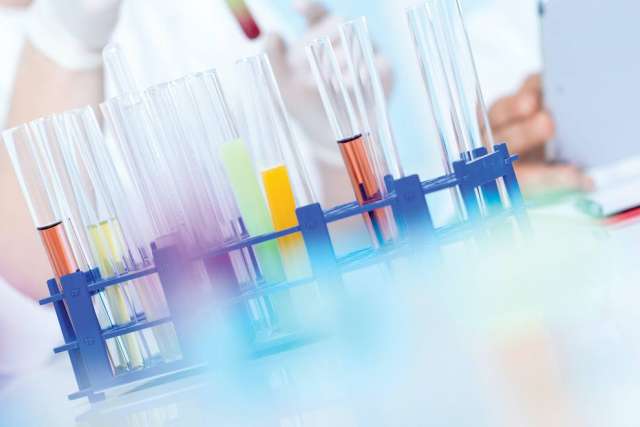 A rack of test tubes containing colorful liquids sits on a lab bench, with a scientist in a white coat visible but blurred in the background.
