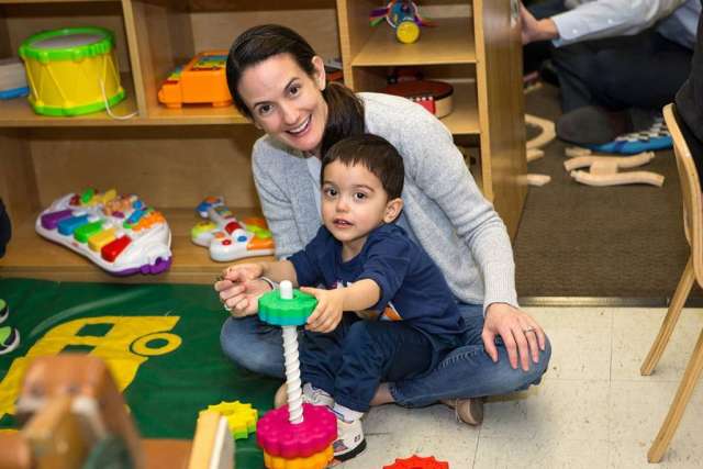 A smiling woman with long brown hair and a young child kneel on the floor in a playroom surrounded by toys. The child plays with a stacking toy.