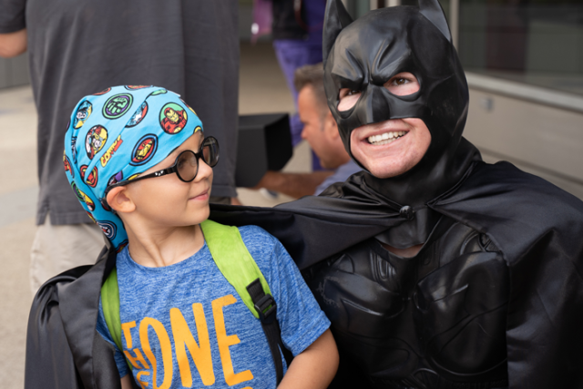 Young UCLA Health patient smiles beside a costumed Batman window washer during a superhero visit at the hospital.