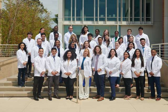 Group of people in white coats pose for a photo outside a modern building with glass windows and stairs.