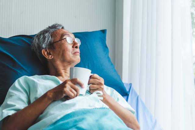 Person lying in a hospital bed, wearing a light blue gown and holding a white mug near a window with sheer curtains.