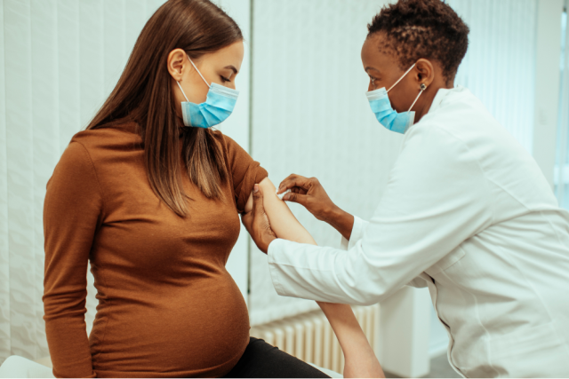 a pregnant patient wearing a mask, receiving a vaccination from a medical professional who is wearing a mask and a lab coat
