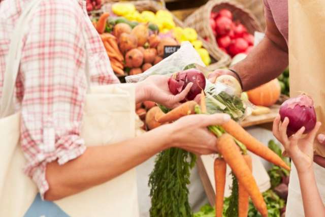 People exchanging fresh vegetables like carrots and onions at a farmers market with baskets of produce in the background.