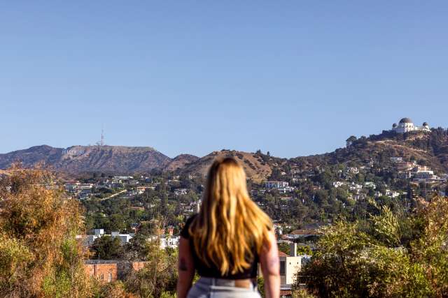A woman, Julie Fordyce, stands with her back to the camera, looking at the Hollywood sign and the Griffith Observatory in Los Angeles.