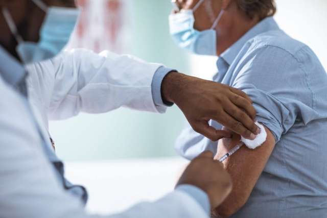 Masked healthcare worker gives a vaccine shot to a masked man in a blue shirt, holding cotton on his arm. 
