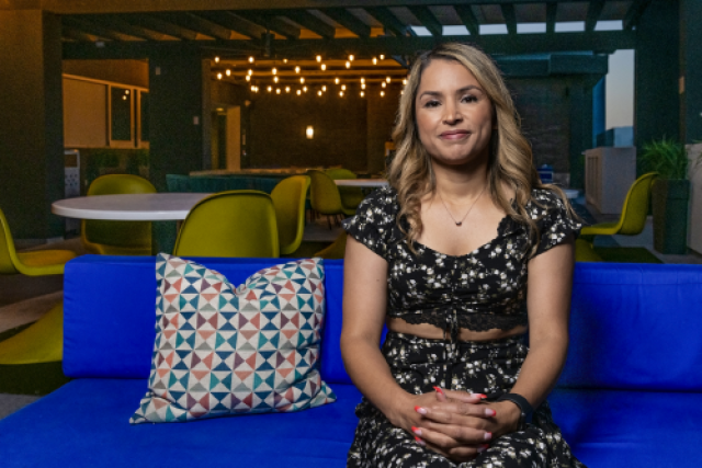 Yadira Ortega seated on a blue couch with a geometric pillow, smiling in an outdoor lounge area with string lights and modern decor.