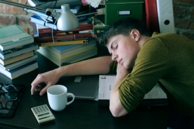  A young person sleeping face down on a desk cluttered with books, a lamp, a laptop, a mug, and a calculator.