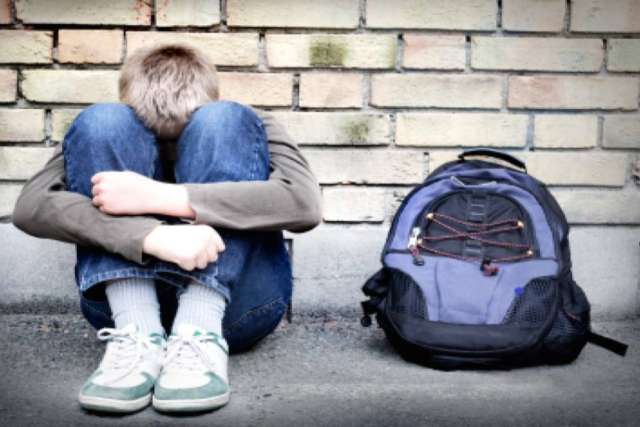 A boy sitting against a brick wall, hugging his knees, with a backpack beside him.