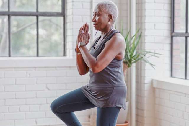 Person practicing yoga indoors, standing on one leg with hands together in front of chest near large windows and a potted plant.