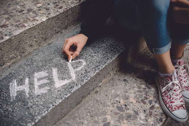 A close-up of a person sitting on rough steps, writing the word "HELP" in white chalk on the stone surface.