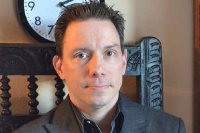 A man in a suit sitting near a clock on the wall.