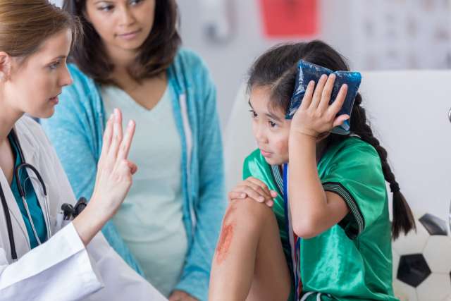 Doctor tests vision of girl holding ice pack to her head as her mother looks on. 