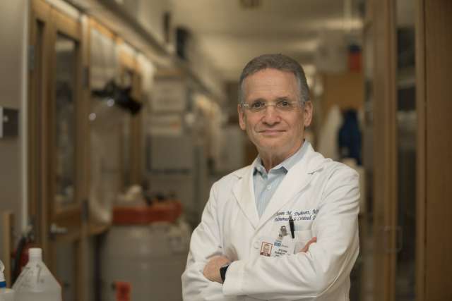 A portrait of Steven M. Dubinett, MD, with glasses and short gray hair, wearing a white lab coat with his name and credentials visible, standing with his arms crossed in a medical or research setting.
