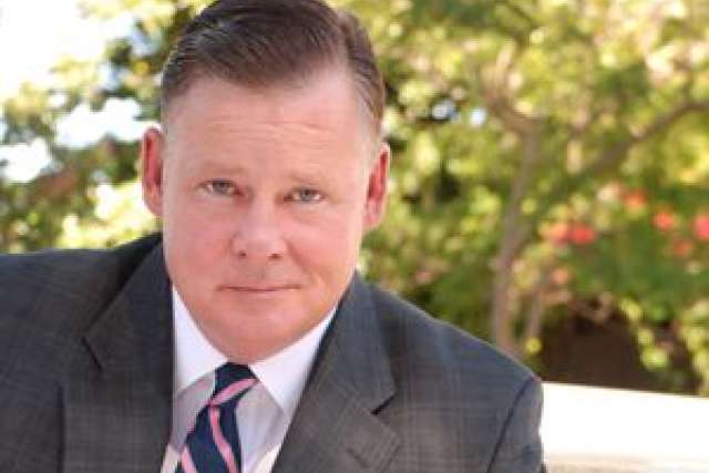 Portrait of Joel Murray in a suit, seated outdoors with greenery behind.