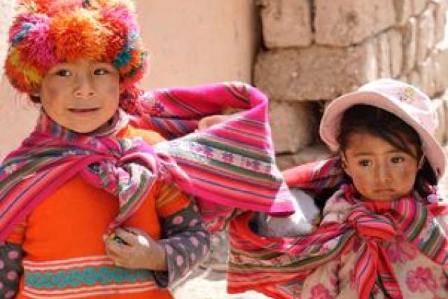 Two Latino children wearing traditional clothing and colorful headwear in a rural setting.