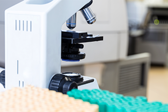 A close-up of a white microscope in a laboratory, with yellow and green racks of test tubes in the foreground.