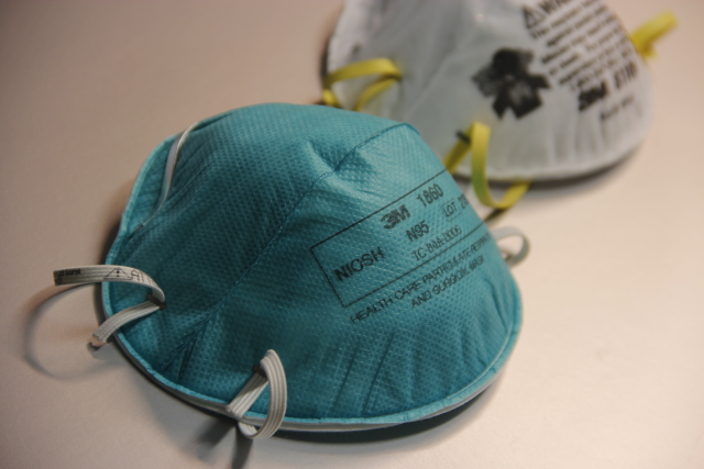 Close-up of stacked N95 respirators with elastic straps, displayed on a table with a blurred background.