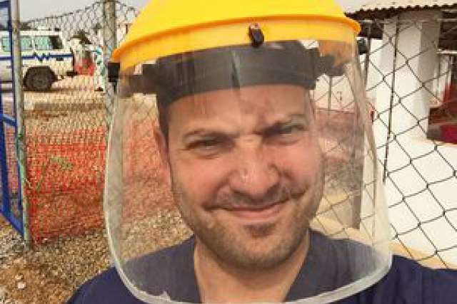 A person wearing a face shield and yellow hard hat, in an outdoor medical setting.