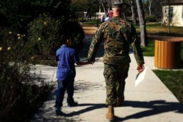 Soldier in military uniform walks hand-in-hand with a child down a path.