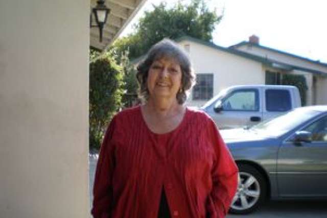 Older woman wearing a red cardigan stands outdoors near a house and car.